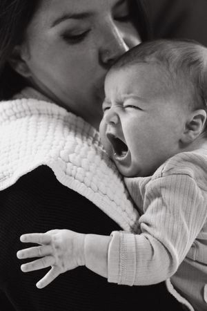 Black and white photo of a crying baby in a striped outfit being comforted against a white knit sweater.