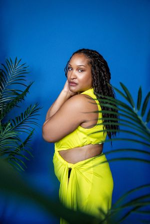 Person in vibrant yellow dress poses against blue backdrop with tropical palm fronds in foreground.