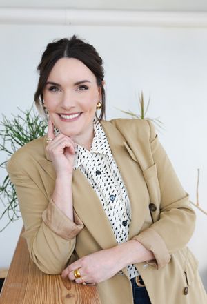Professional portrait in tan blazer and patterned top against white background.