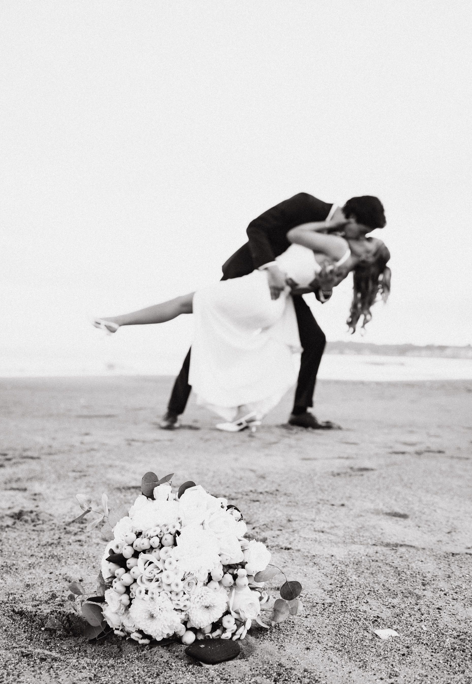 Black and white beach wedding photo with bouquet in foreground and couple embracing.