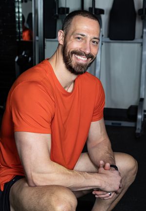 Athletic man in orange t-shirt smiling at camera in gym setting.