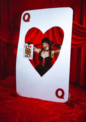 Woman in black corset poses through Queen of Hearts playing card cutout against red draped background.