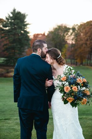 A couple in wedding attire share intimate moments while holding autumn-colored floral bouquets in a park setting.