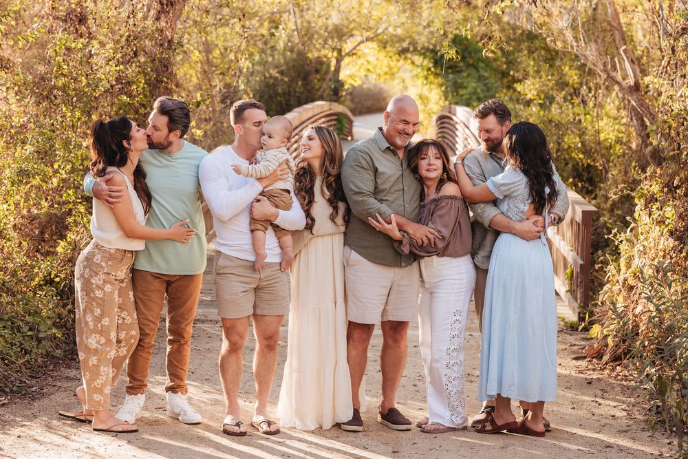 Large family group portrait outdoors on a wooden path, couples embracing among lush greenery in neutral tones.