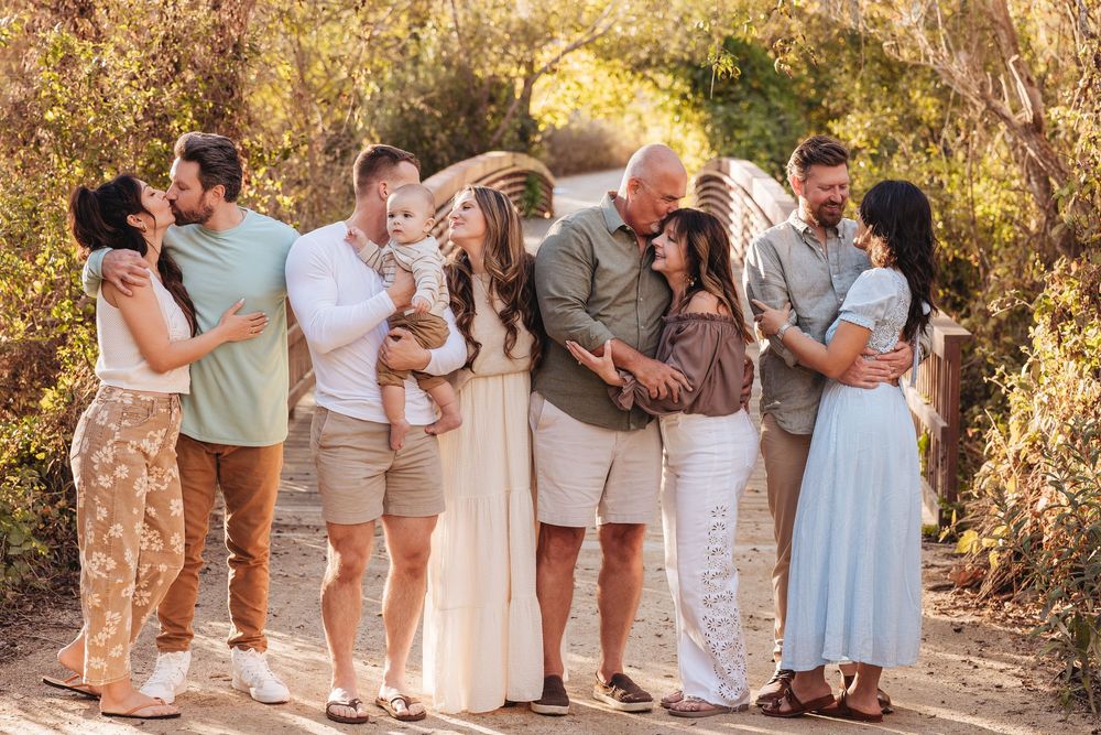 A joyful extended family embraces and kisses on a wooden bridge surrounded by lush greenery during an outdoor photo session.