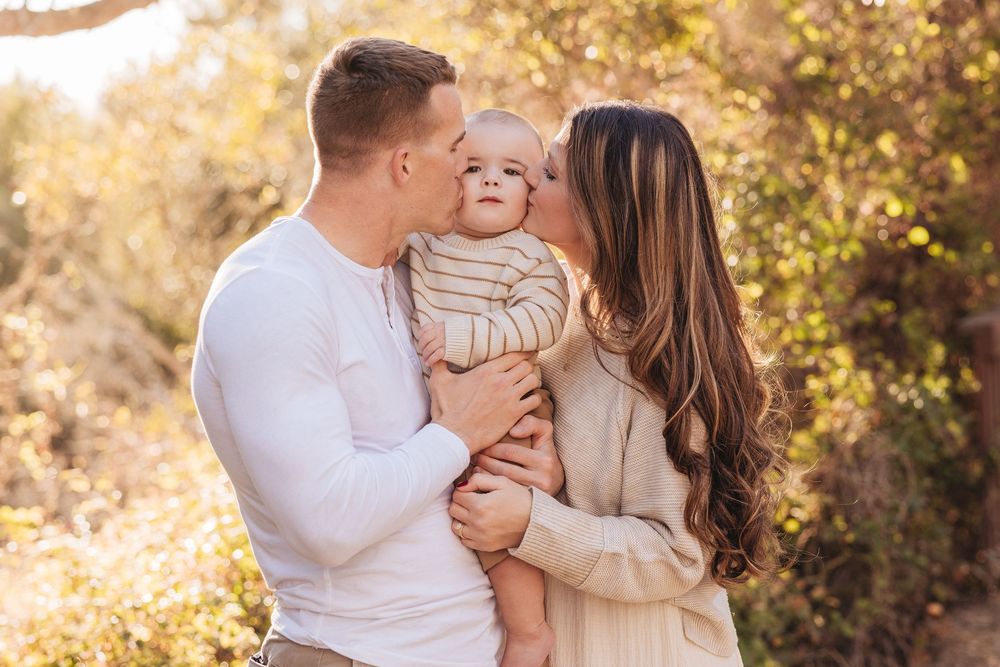 Young couple kissing their baby outdoors in golden sunlight among green foliage.
