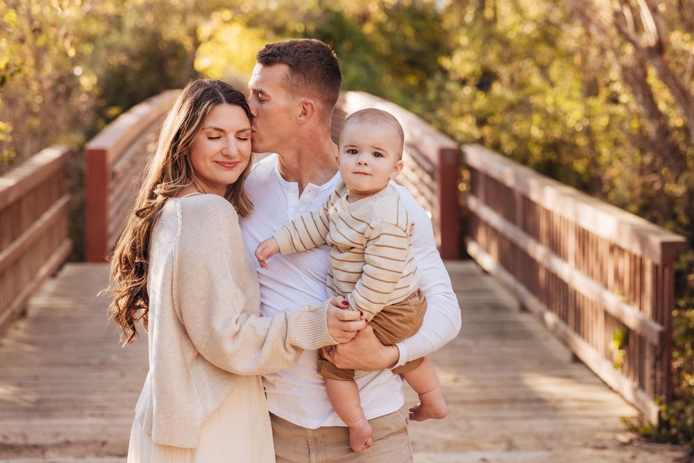 Young family with baby on a wooden bridge, parents smiling and kissing their infant son outdoors.