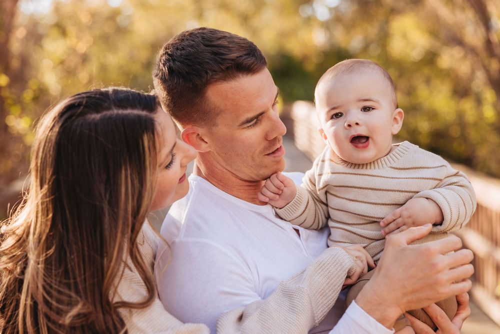 Happy young couple smiling at their laughing baby boy outdoors in warm natural light.