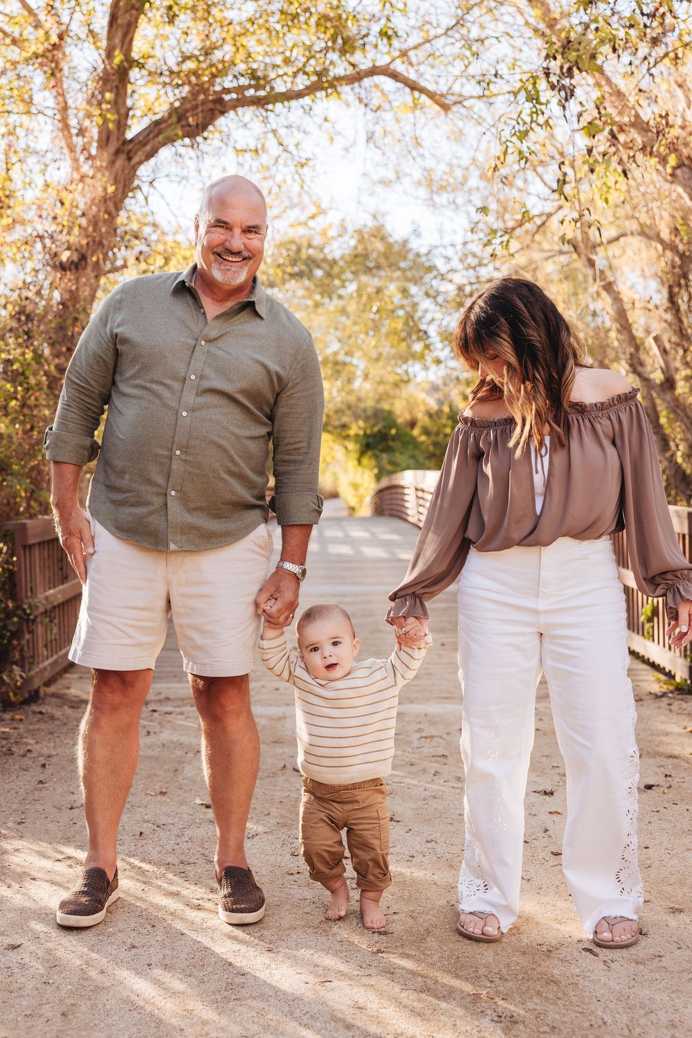 A smiling family with a bald father, brunette mother, and baby toddler walking outdoors on a sunny dirt path.