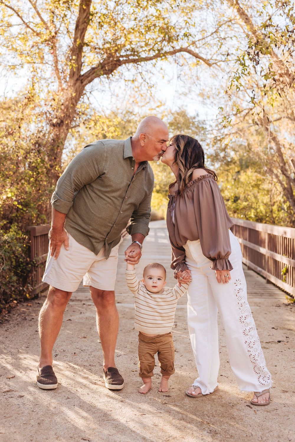 Couple kissing while holding hands with their baby on a sunny outdoor bridge surrounded by autumn trees.