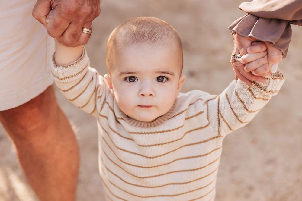 Adorable baby in striped outfit learning to walk while holding adults hands outdoors.