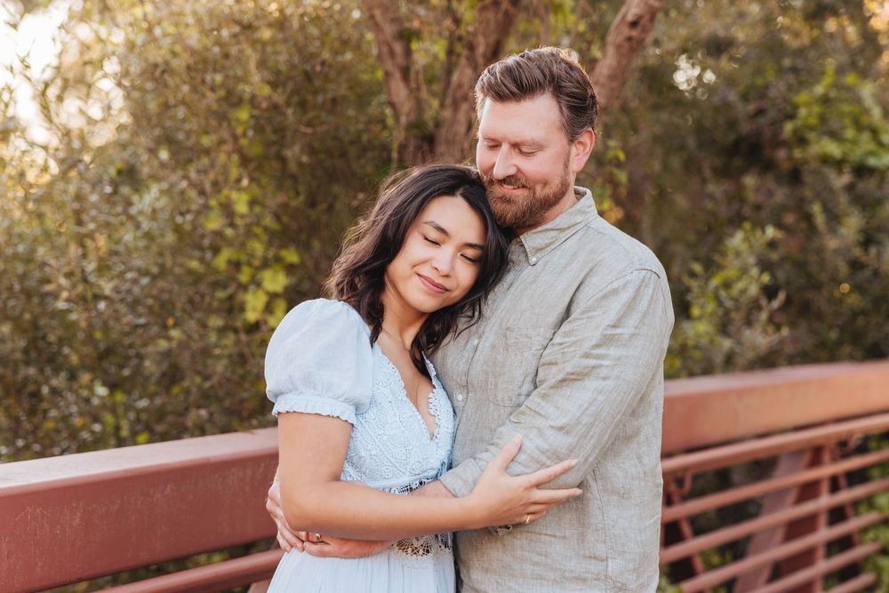 Couple embracing on a bridge surrounded by lush green trees in a warm outdoor engagement photo.