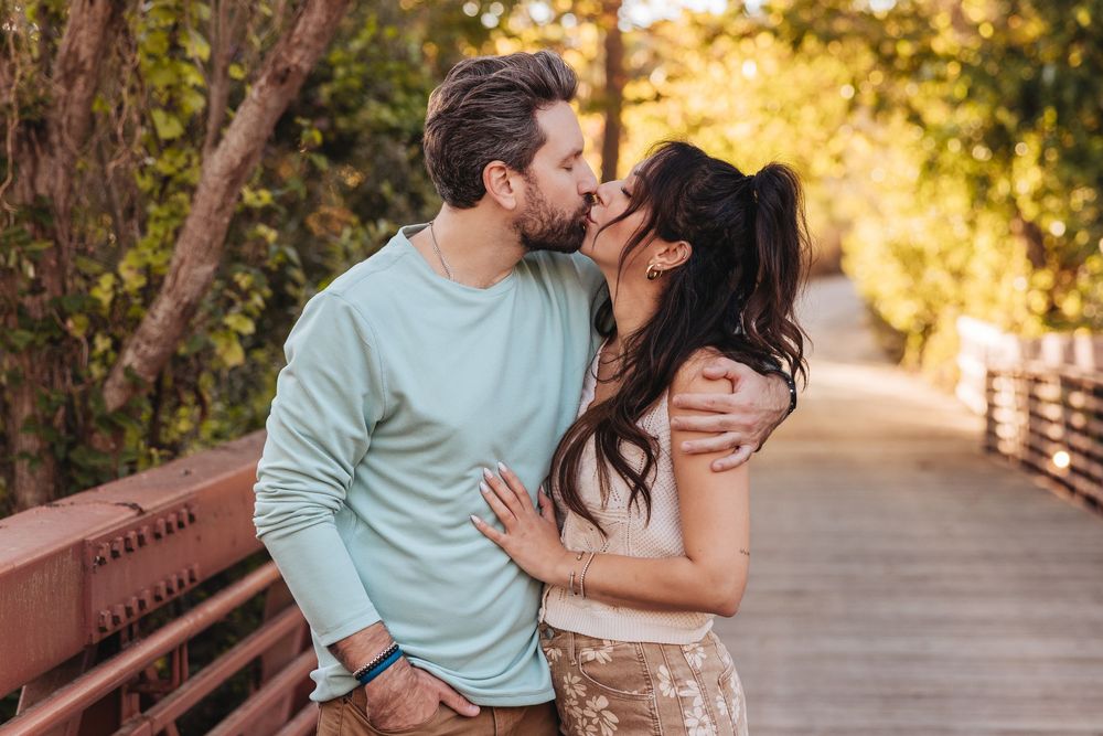 Couple sharing a romantic kiss on a wooden bridge surrounded by autumn trees in a park setting.