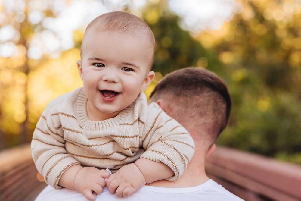 Happy baby in striped sweater laughing while being held by father outdoors in autumn park setting.