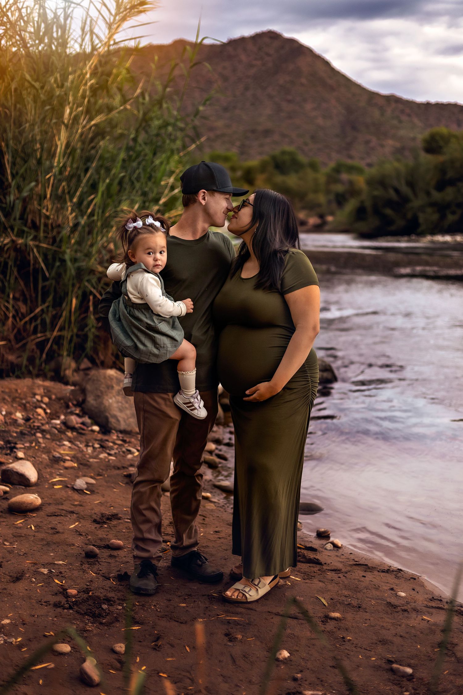 Pregnant woman in floral dress holds hands with toddler girl and man on desert path with saguaro cacti at sunset.