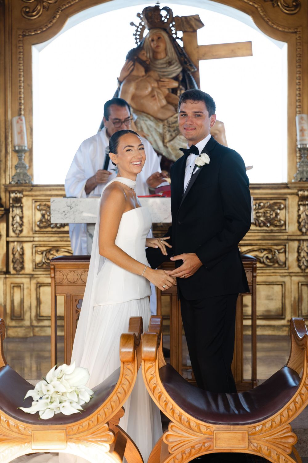 Bride and groom hold hands at the altar of an ornate Mexican church during their wedding ceremony.