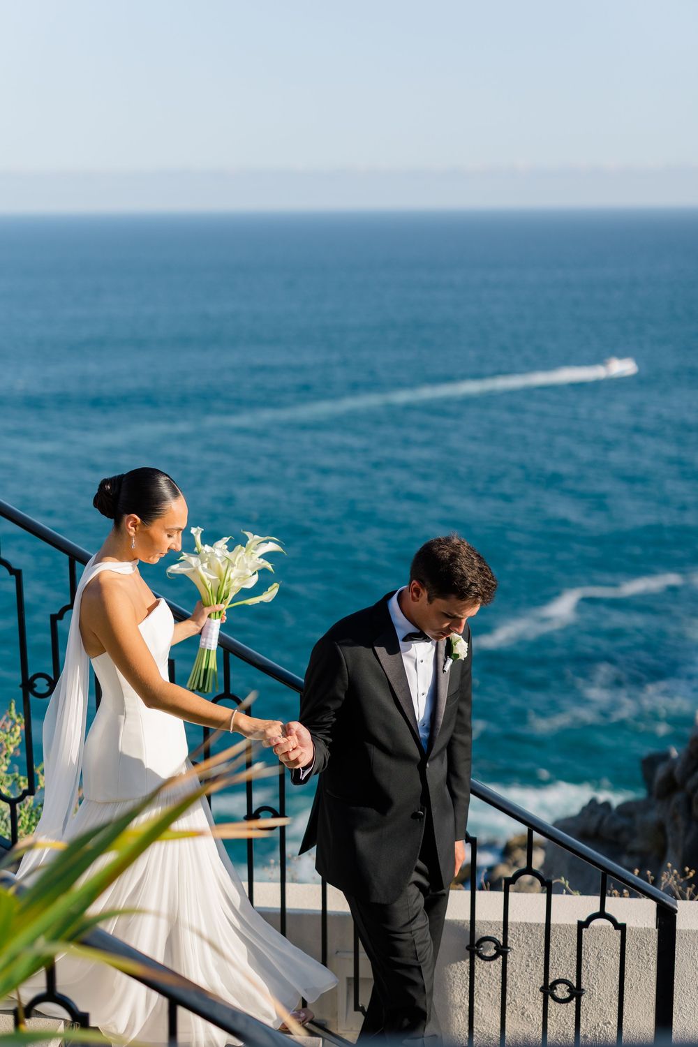 Elegant bride and groom descend iron staircase steps with breathtaking blue ocean backdrop.