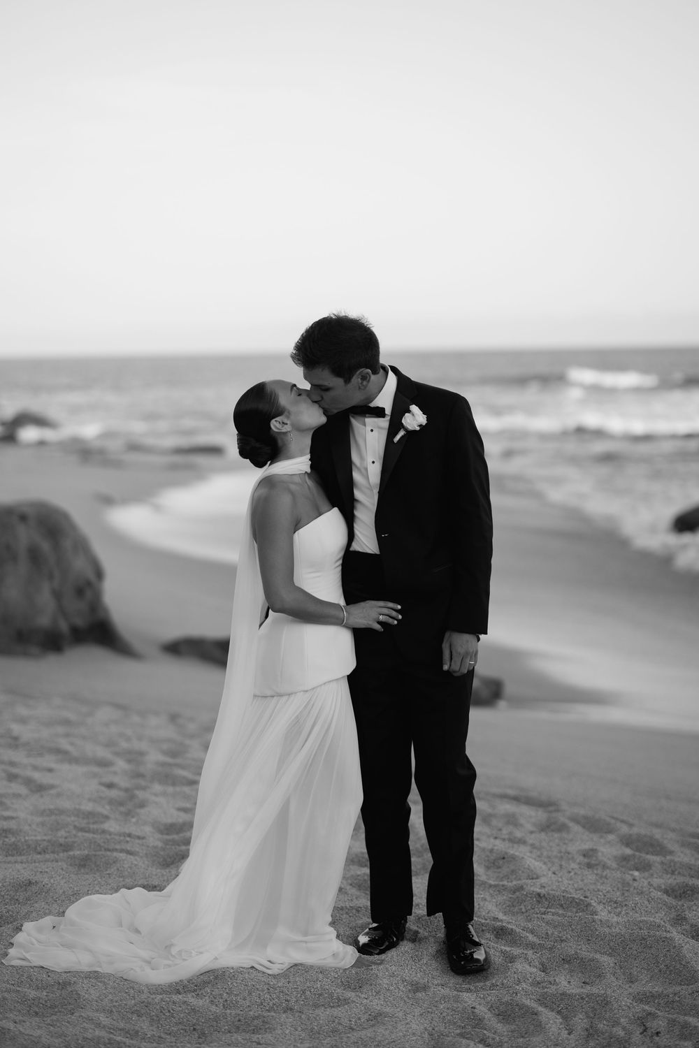 Black and white photo of newlyweds sharing a kiss on a sandy beach at dusk.