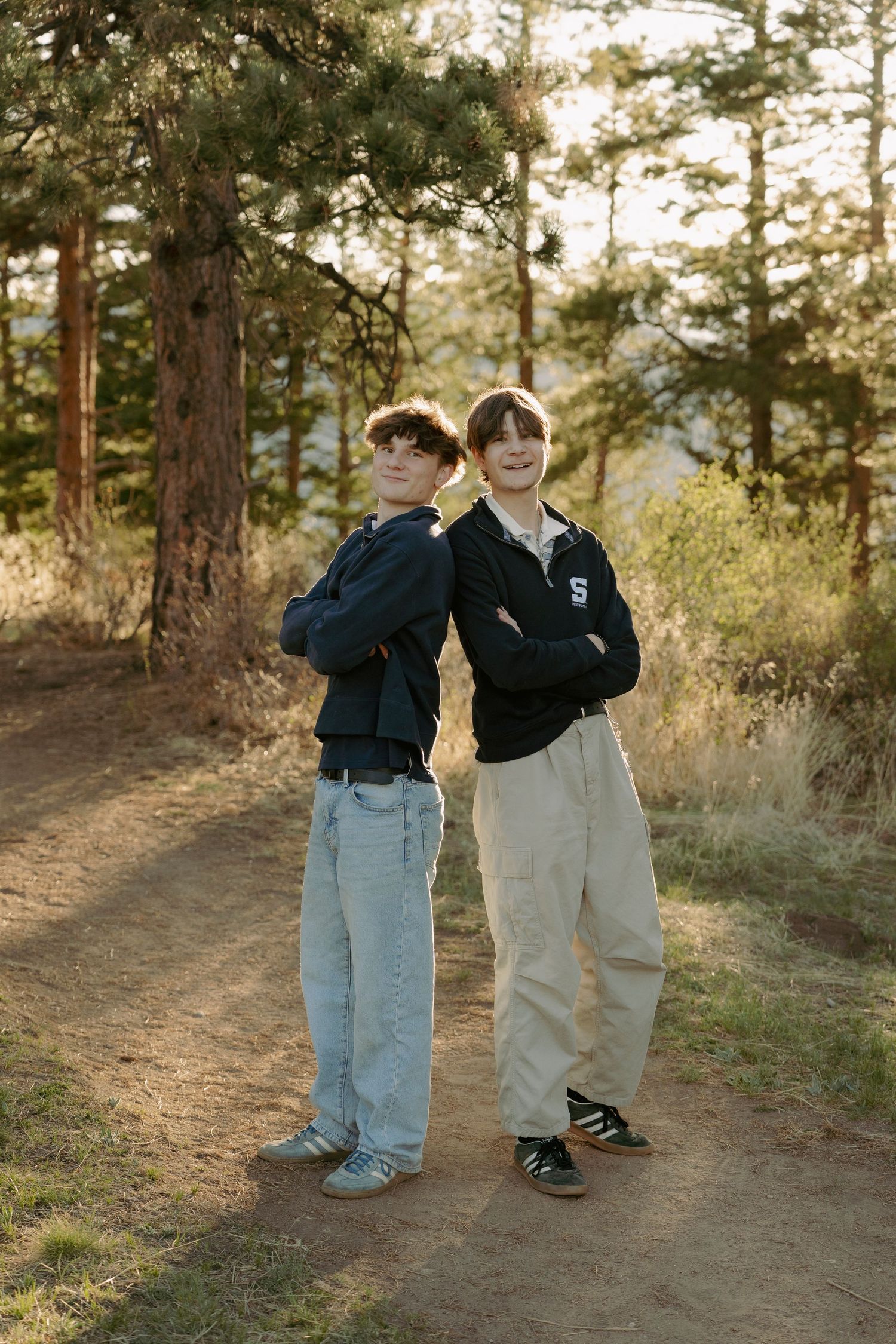 Two teenage boys stand back-to-back with arms crossed on a forest trail, wearing casual streetwear and Adidas shoes.