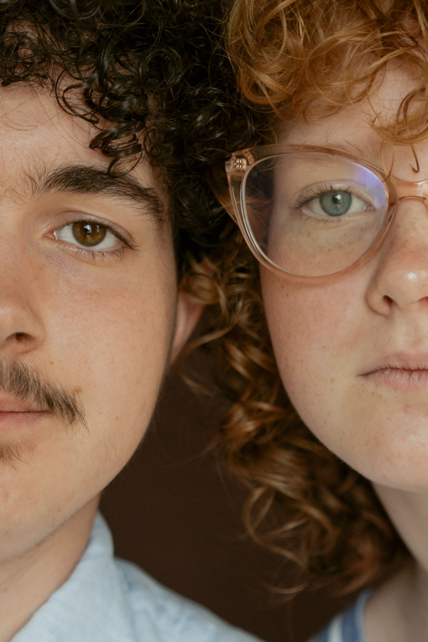 Close-up portrait of two young people with curly hair, one with glasses, posing cheek to cheek.