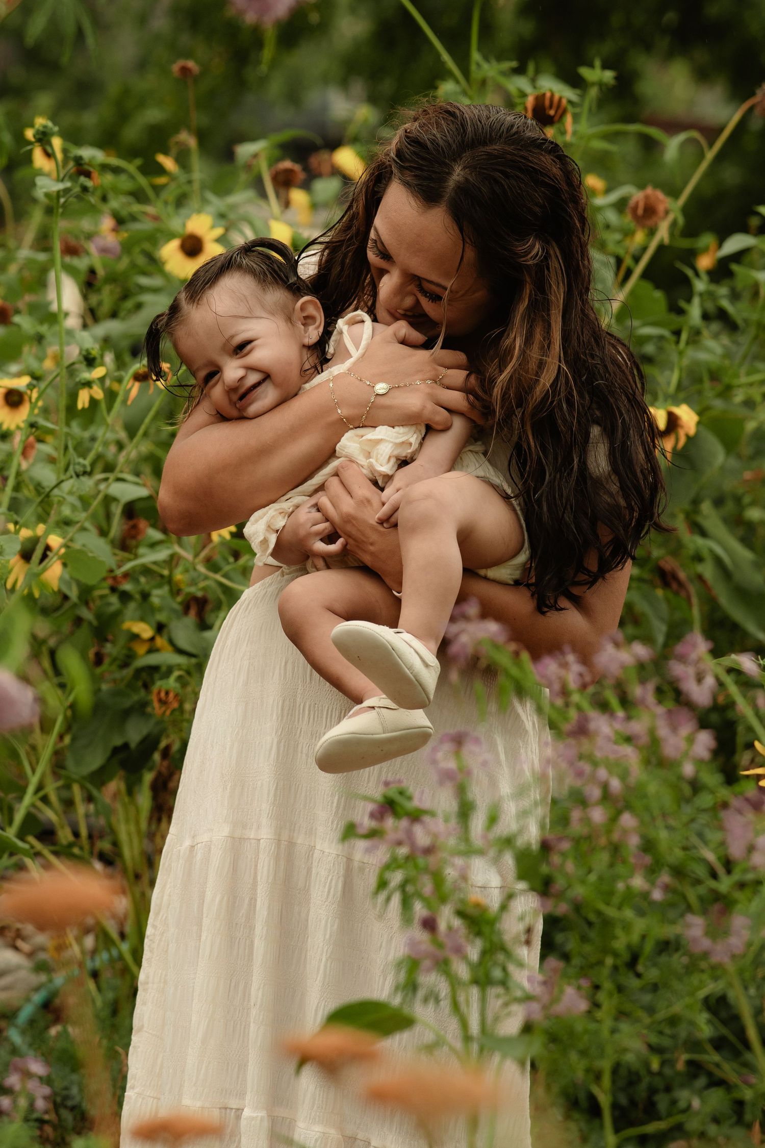 A mother in a white dress joyfully holds two young children in a lush garden filled with colorful wildflowers.