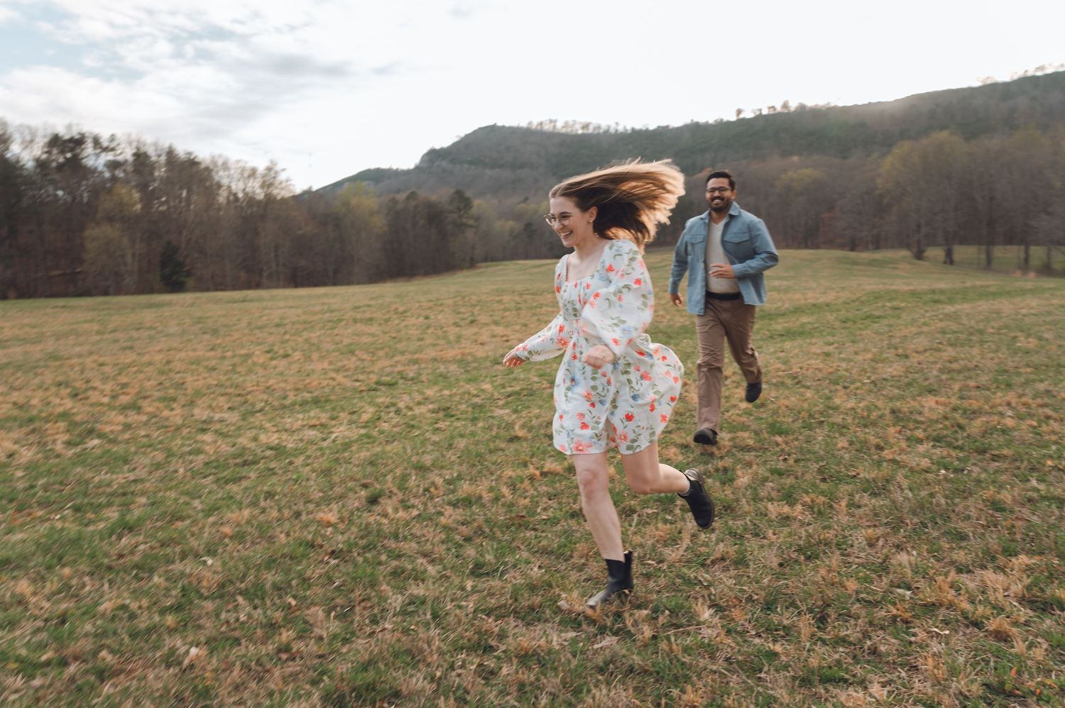 Couple running and laughing in green field with mountain backdrop on cloudy day.