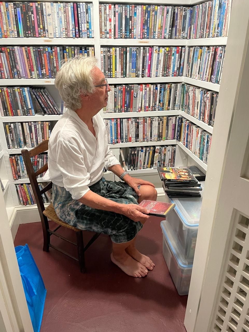 Elderly man sitting in a chair surrounded by floor-to-ceiling shelves filled with DVD and Blu-ray movie collections.