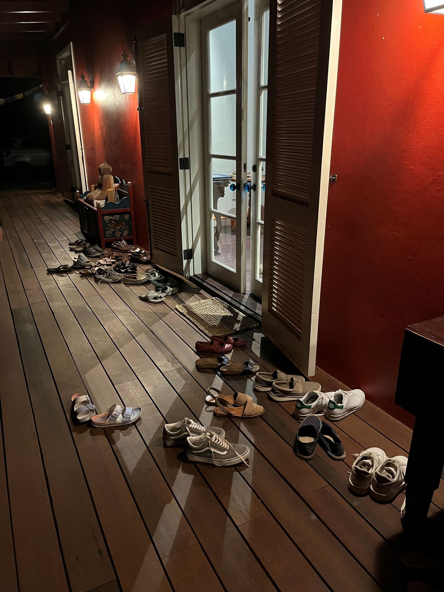 Multiple pairs of shoes lined up on a wooden deck outside a red-walled building entrance at night.