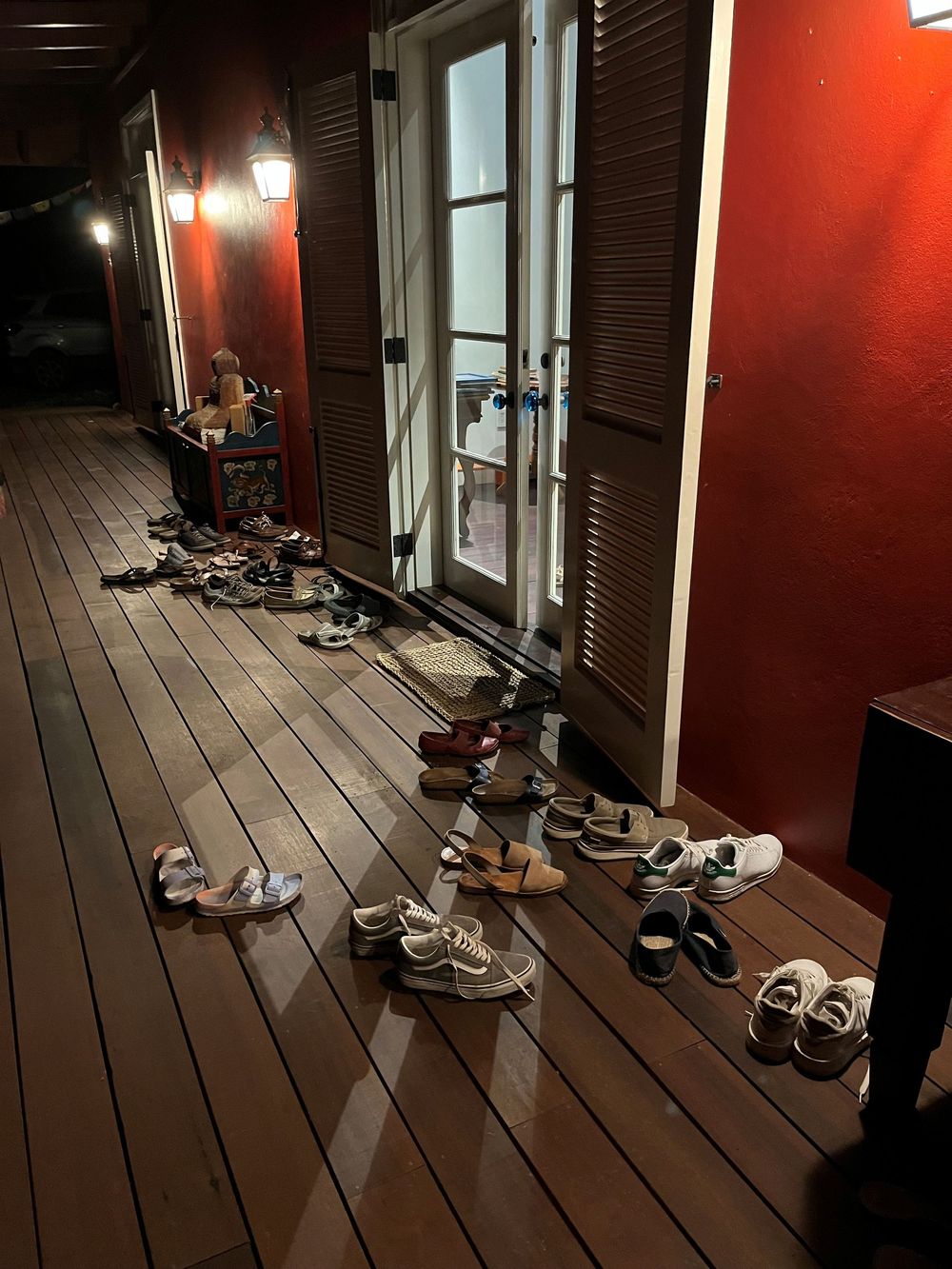 Multiple pairs of shoes lined up on a wooden deck outside a red-walled building entrance at night.