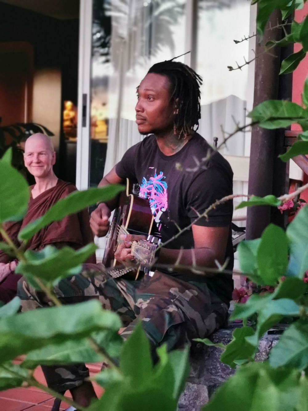 A man with dreadlocks plays acoustic guitar outdoors, wearing a black graphic tee, with a monk seated behind him.