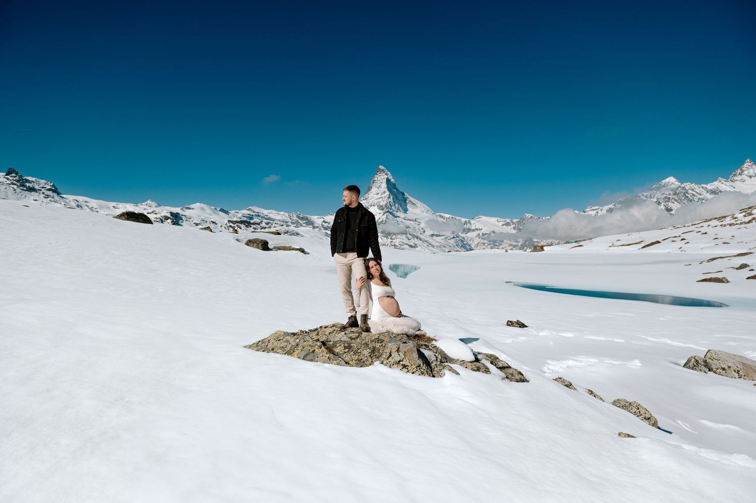 Couple standing on snowy rocks with the iconic Matterhorn peak and frozen alpine lake in Zermatt, Switzerland.