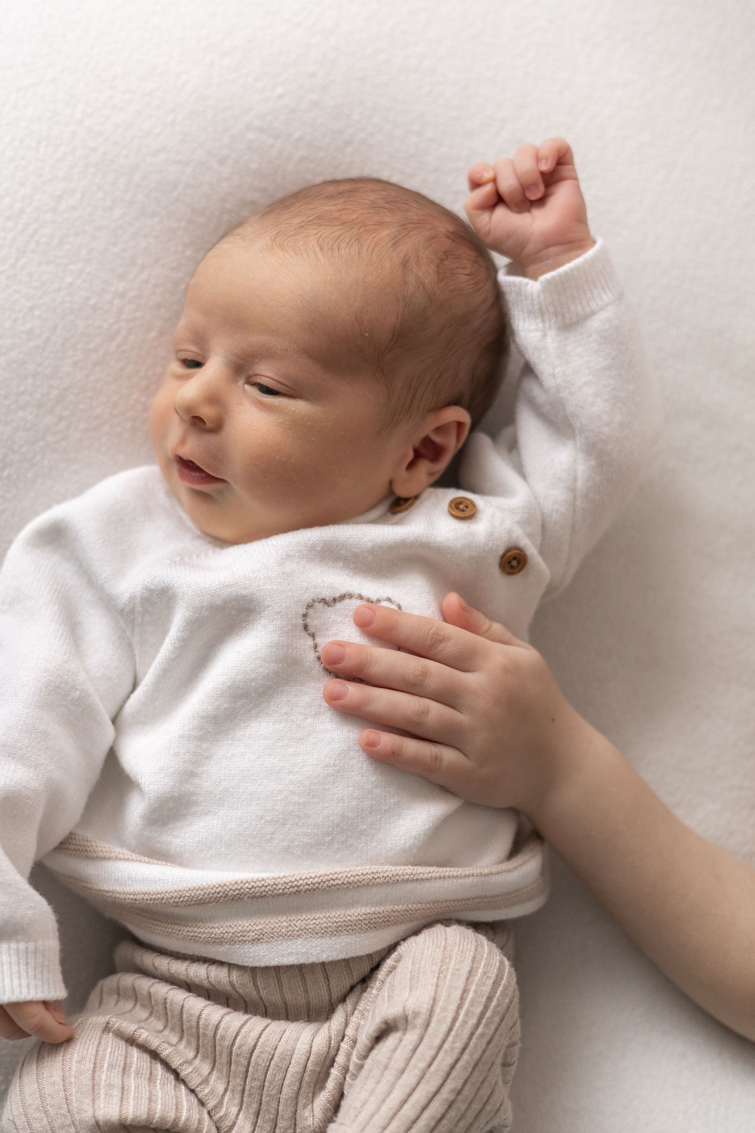 Newborn baby in white onesie and beige leggings lying on white blanket with siblings hand during newborn photoshoot Nottingham