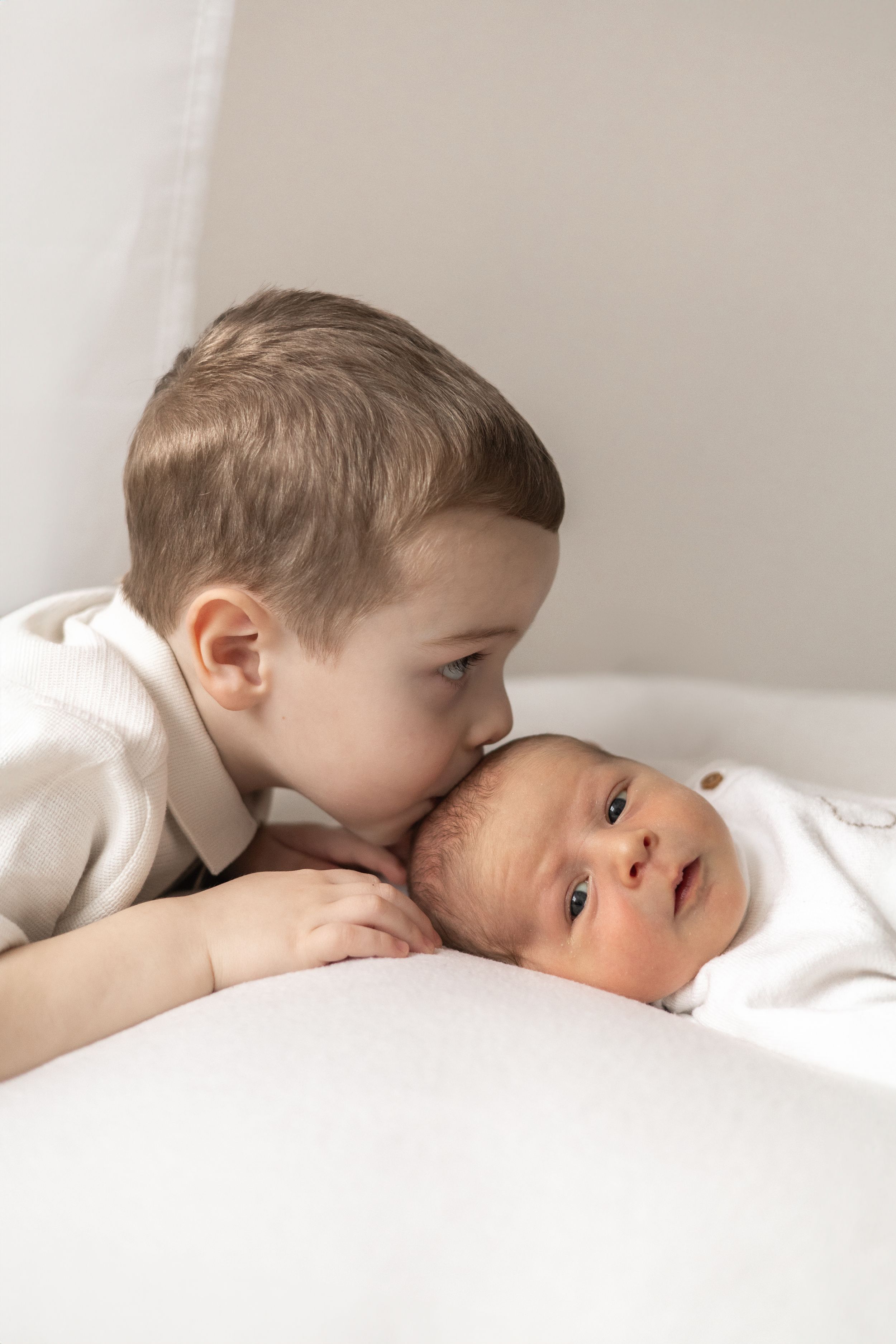 A young sibling gently kisses a newborn baby lying on a white surface, capturing a tender sibling moment newborn photoshoot Nottingham