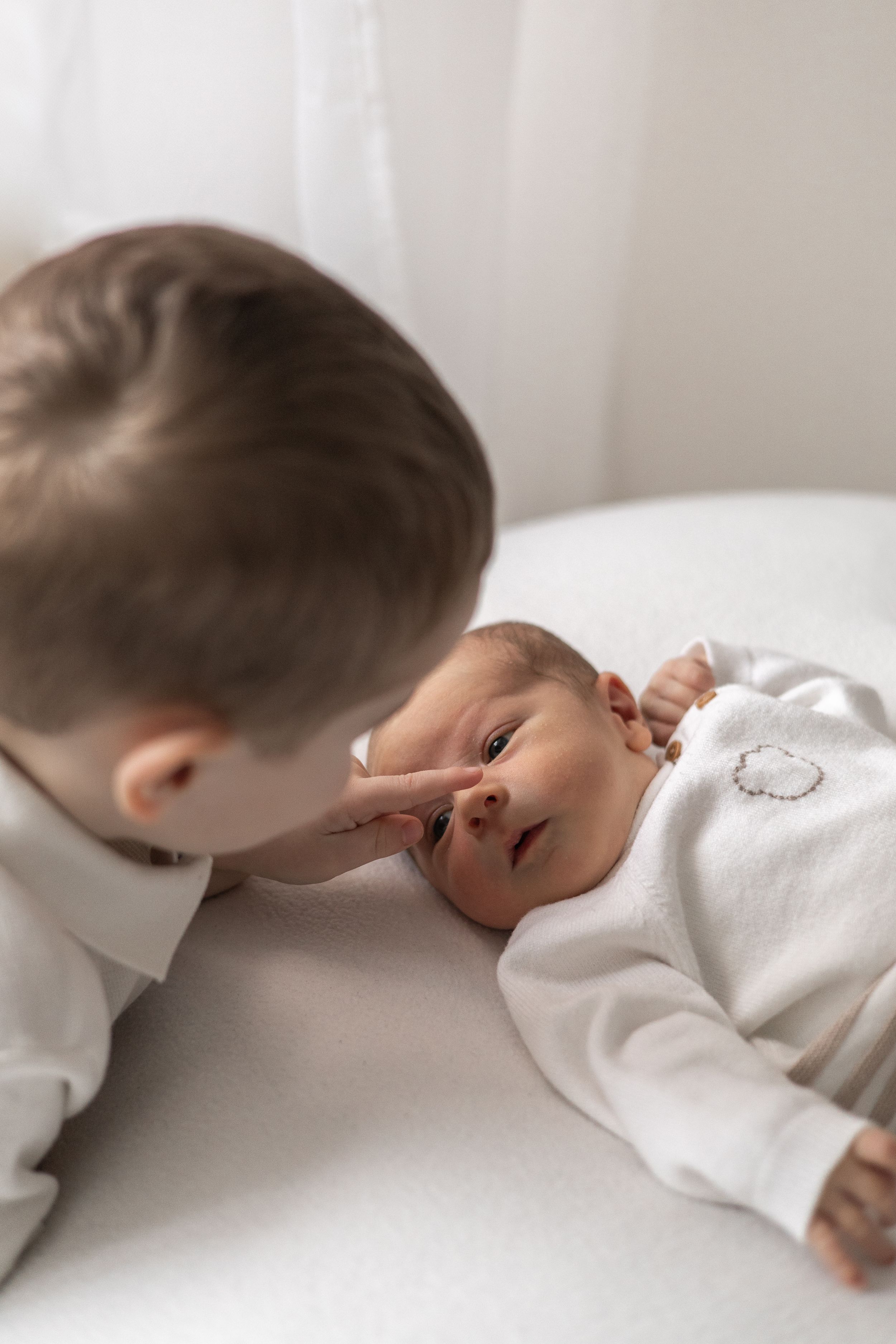 An older sibling gently touches a newborn baby's nose while the baby lies on a white surface wearing a white onesie newborn Nottingham