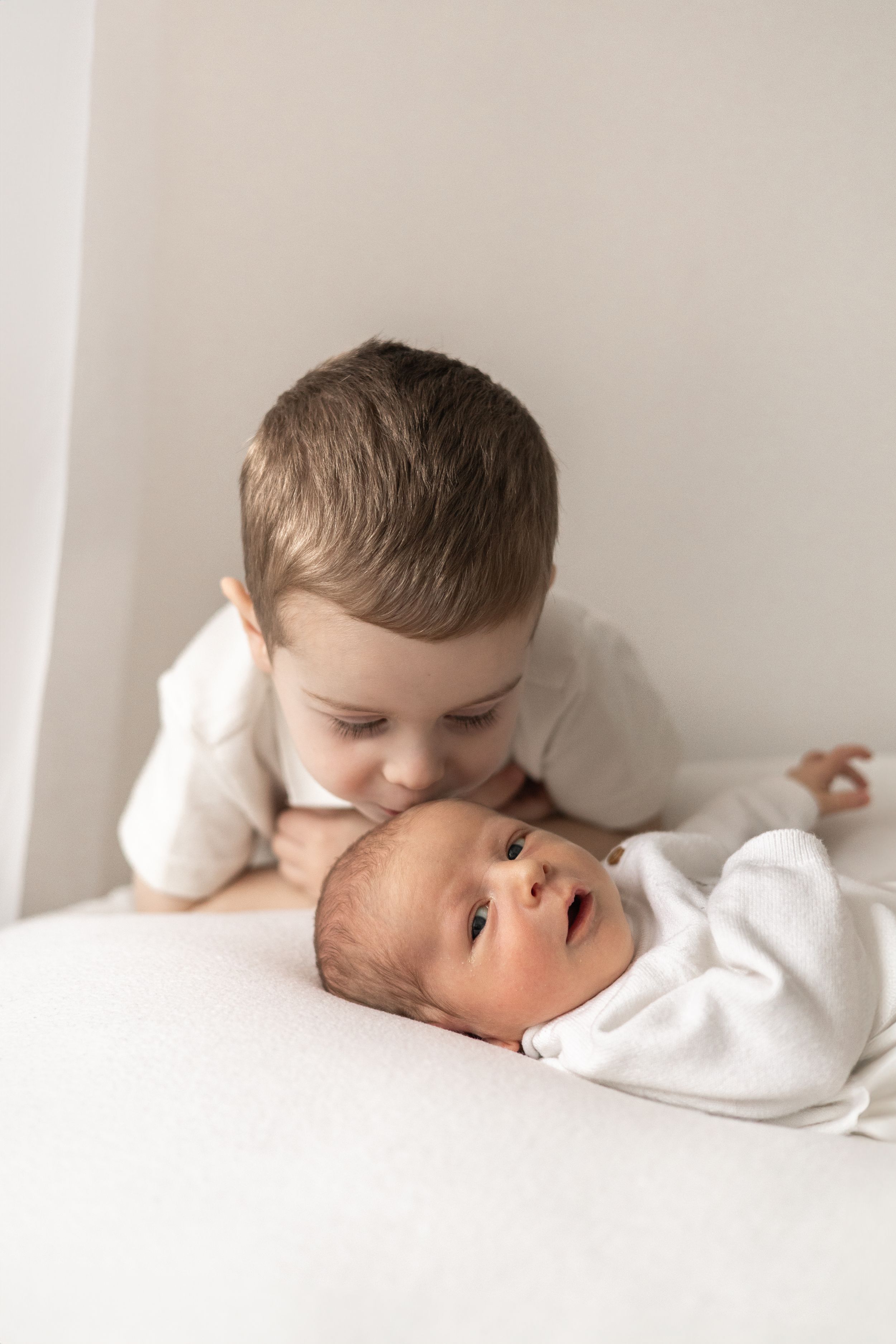 Young sibling in white shirt gently kissing newborn sibling wrapped in white blanket on soft white surface newborn photoshoot Nottingham