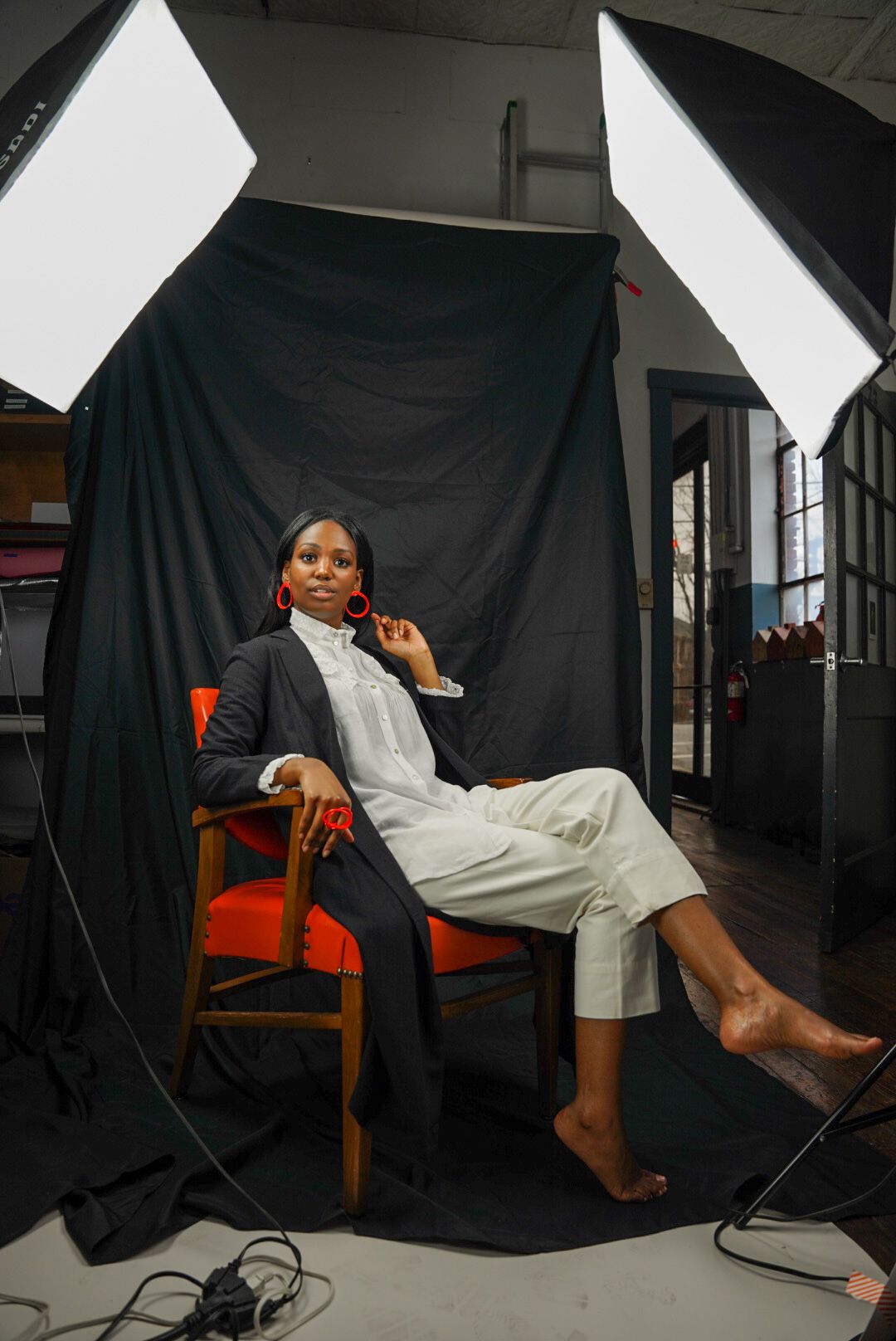 Woman in blazer and white shirt sits on orange chair in professional photo studio with softbox lights.