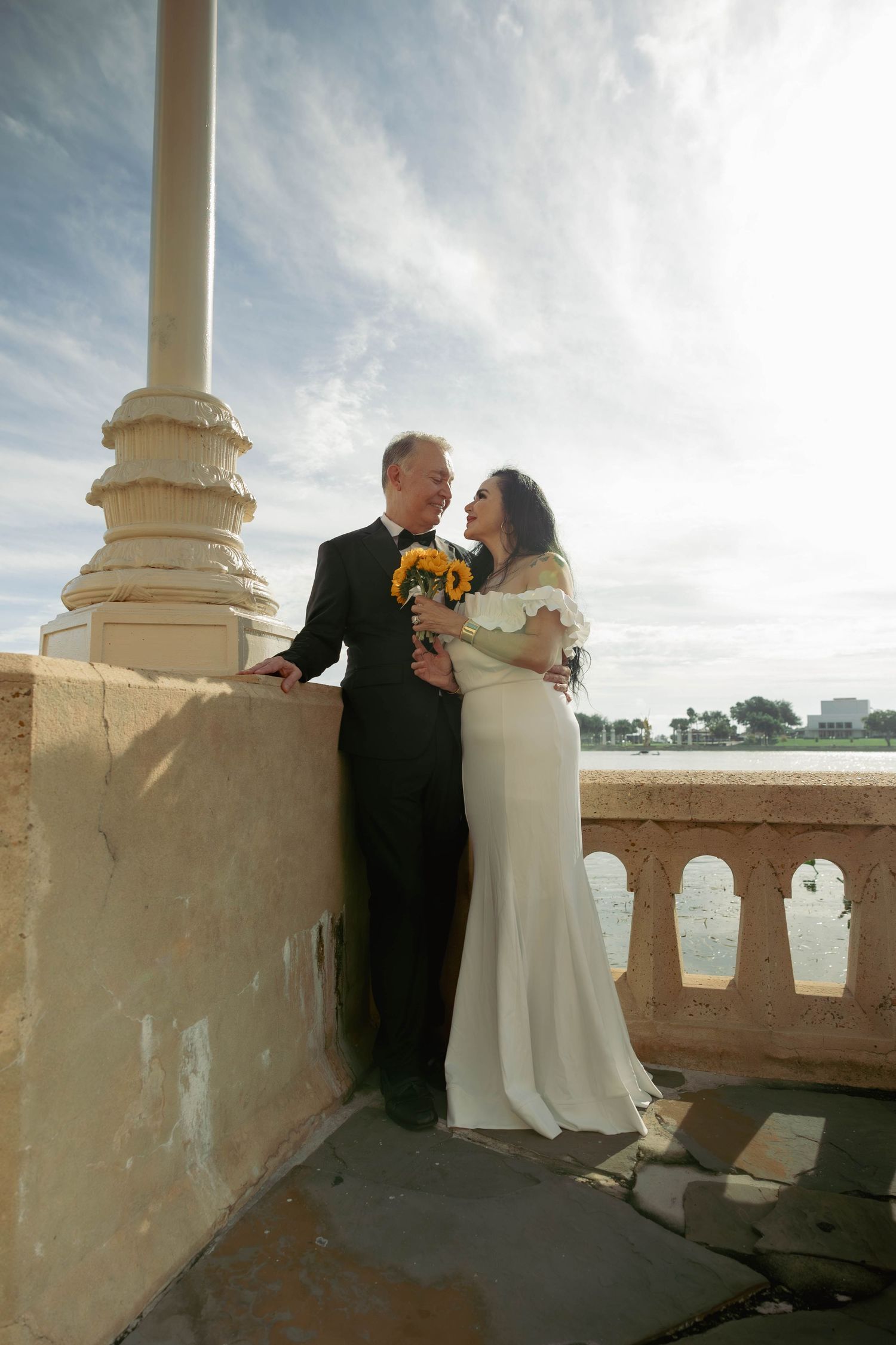 Bride and groom sharing a romantic moment on a waterfront bridge holding sunflower bouquet.