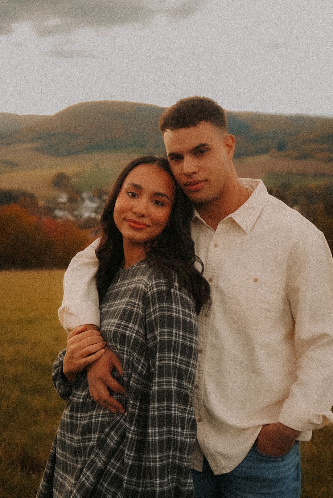 A young couple poses in a scenic autumn field with rolling hills, the woman in plaid and man in a white shirt.