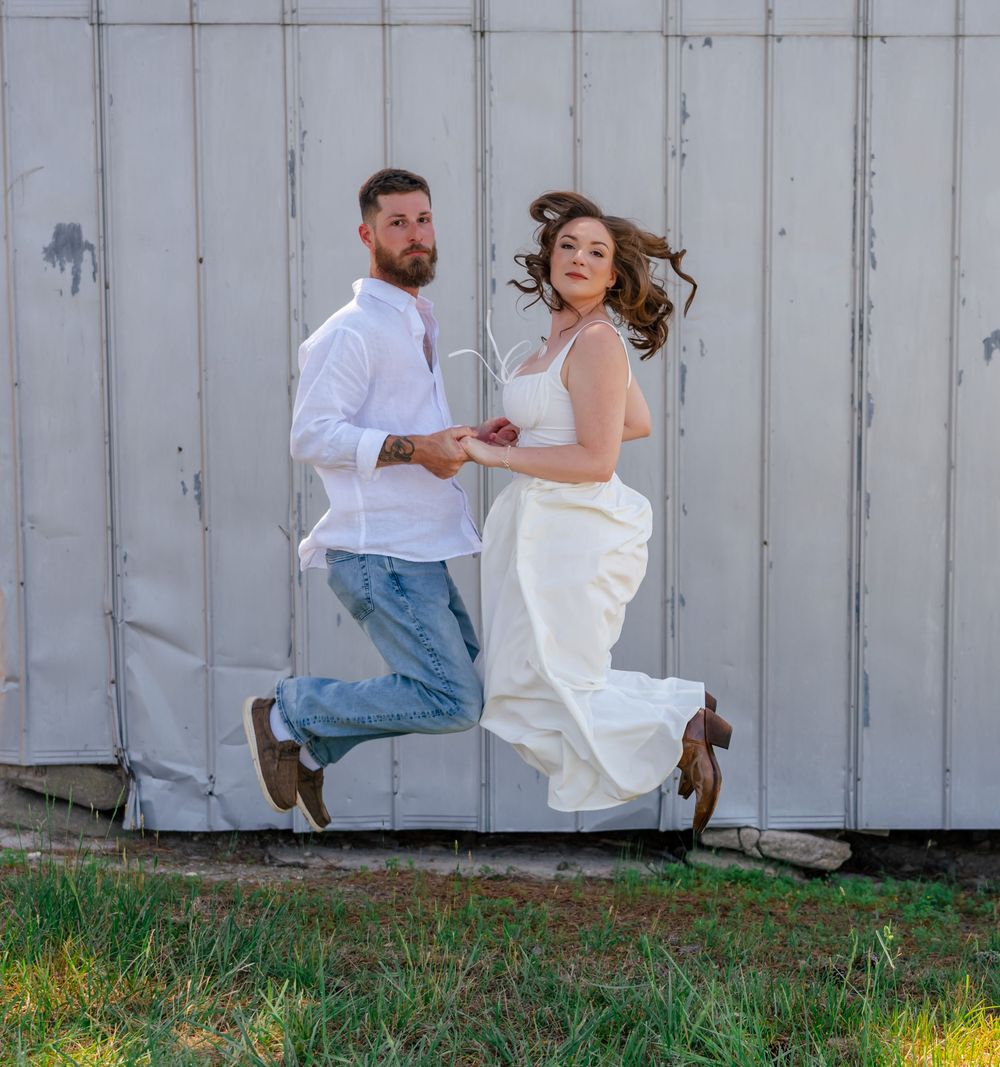 Joyful couple in white outfits jumping in front of a rustic barn wall.