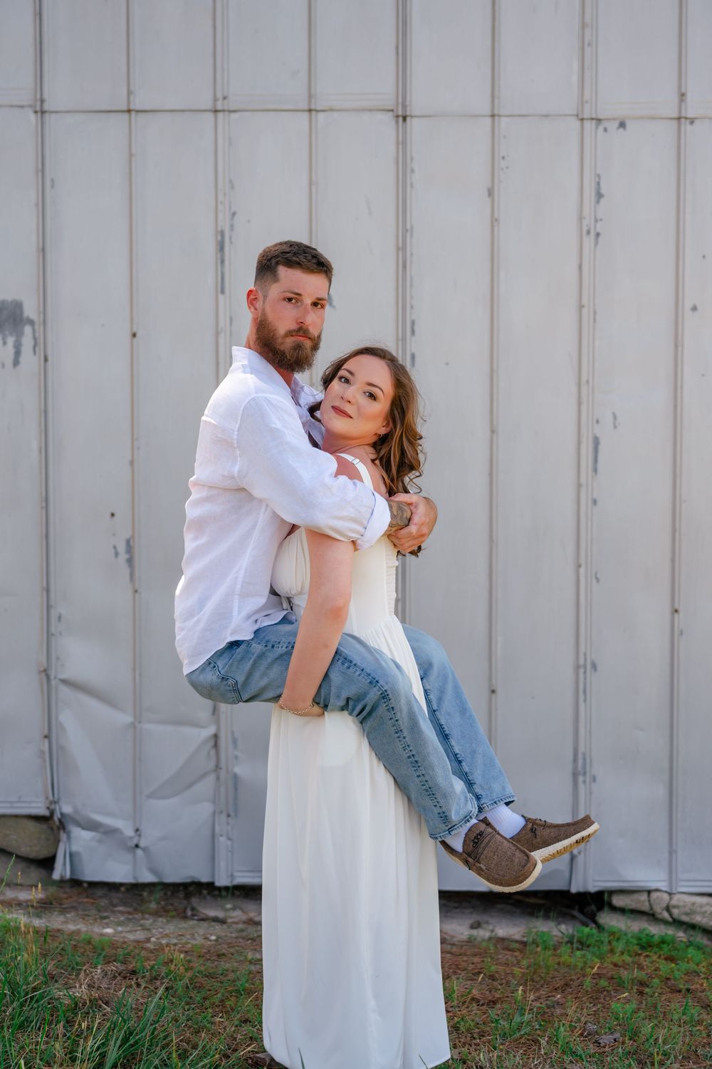 A bearded man in a white shirt and jeans being carried by a woman in a white dress outdoors near a barn.