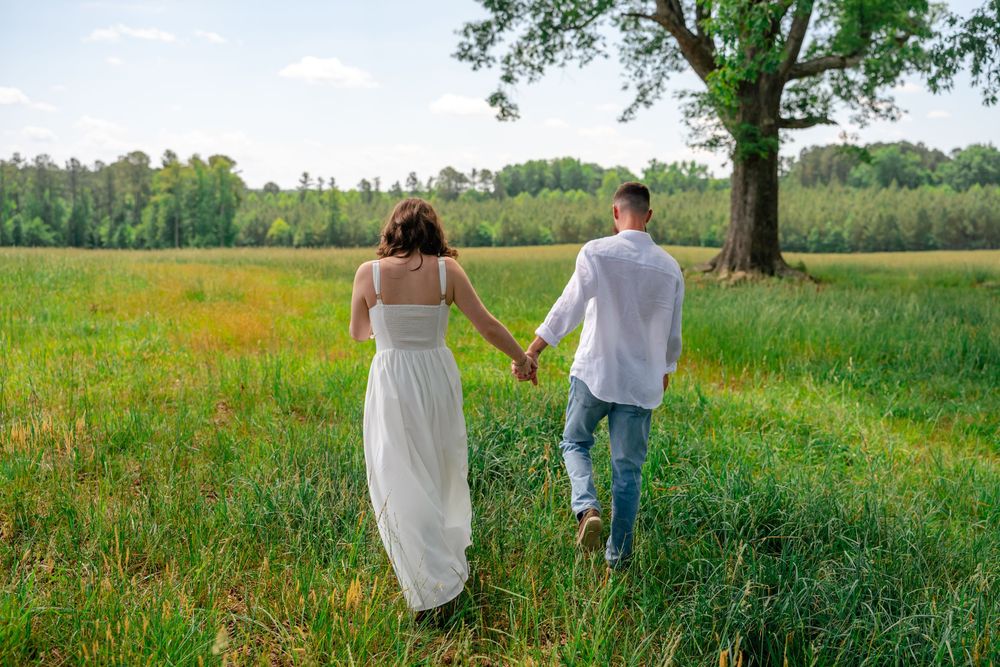 Couple holding hands walking through a lush green meadow near a large tree on a sunny day.