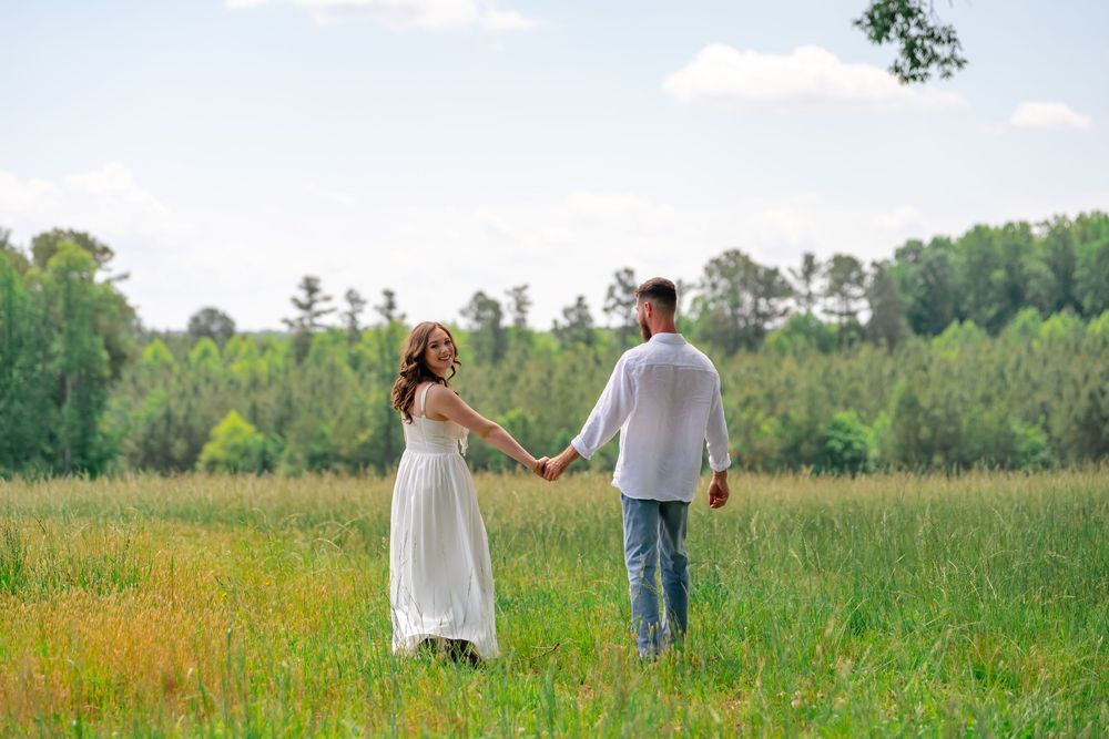 A couple holds hands while walking through a lush green meadow with a forest backdrop on a sunny day.