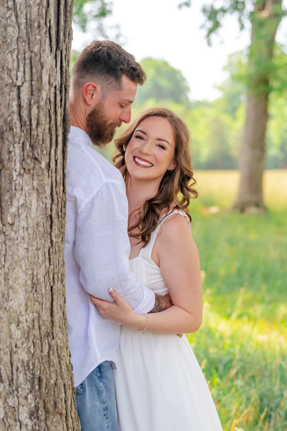 Smiling couple in white outfits embracing by a tree in a sunny green field.