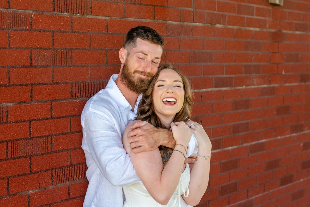 A smiling couple embraces against a red brick wall, the bearded man hugging the laughing woman from behind outdoors.
