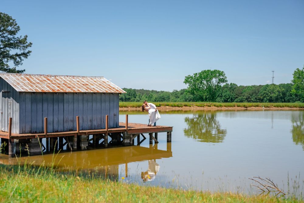 Couple embracing on a wooden dock beside a rustic boathouse overlooking a calm reflective pond in a rural landscape.