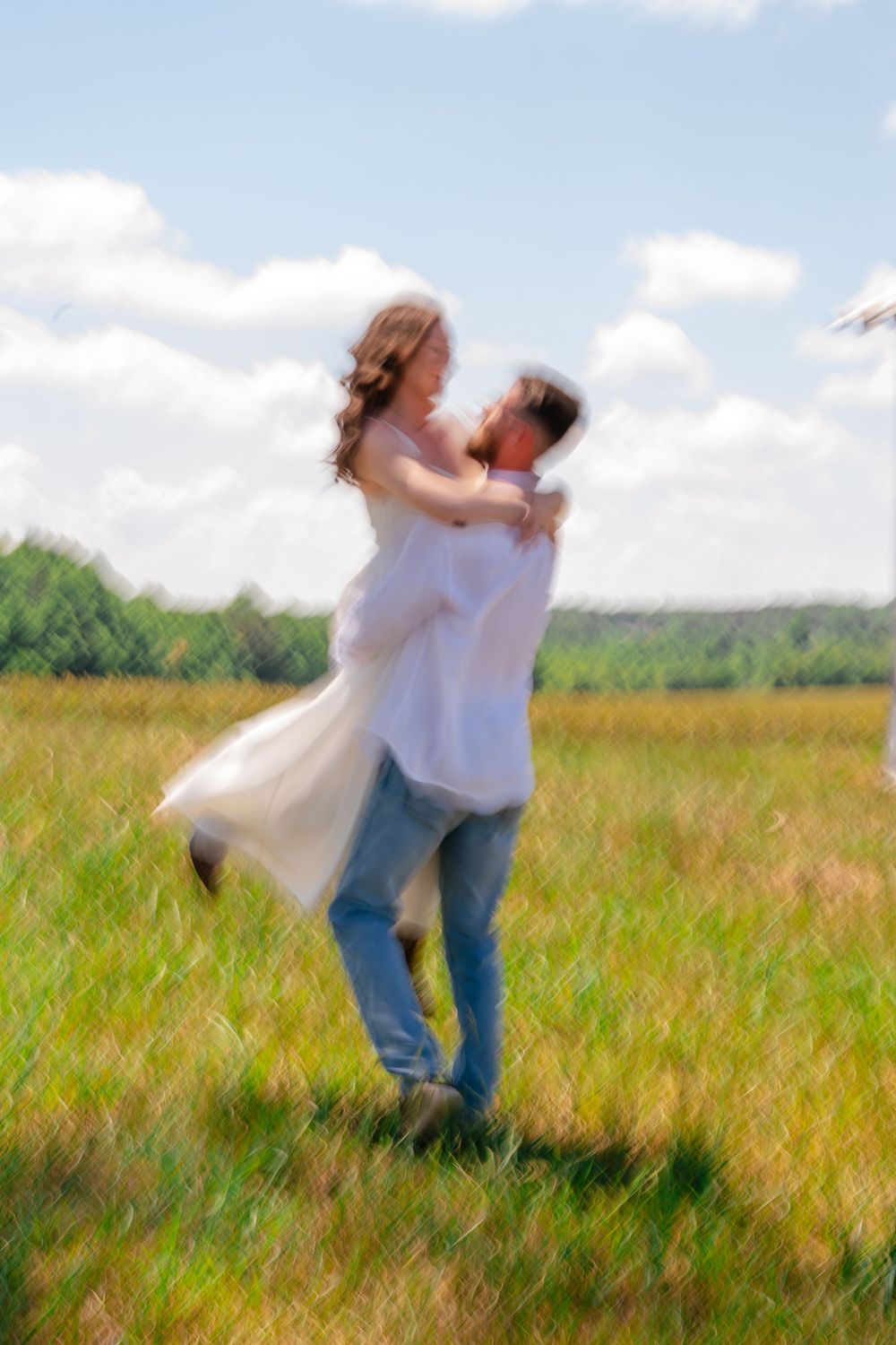 A joyful couple embraces in a sunny green field, man lifting woman in white dress, with blue sky and trees behind them.