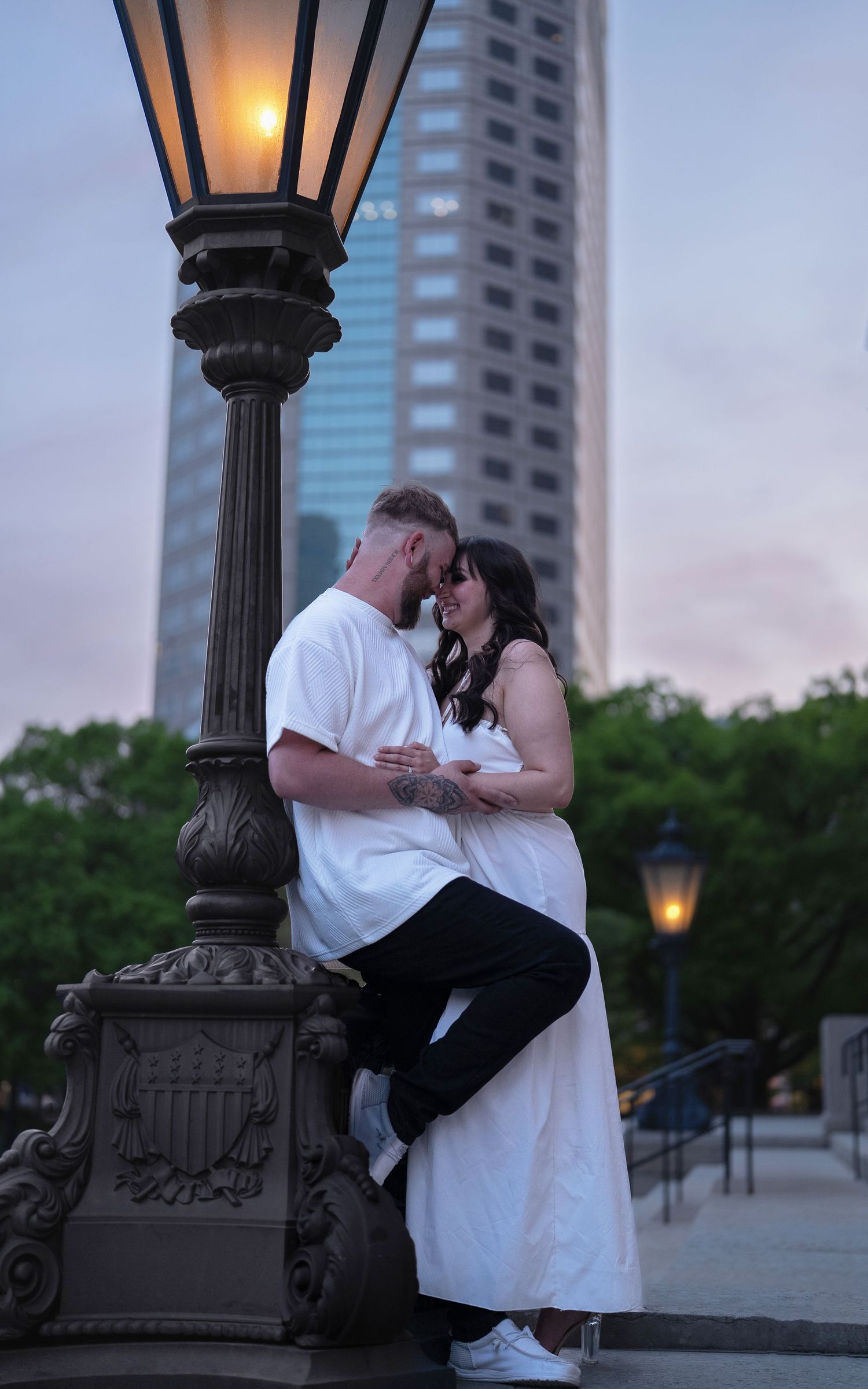 Couple embracing romantically near an ornate street lamp at dusk with a city skyline in the background.
