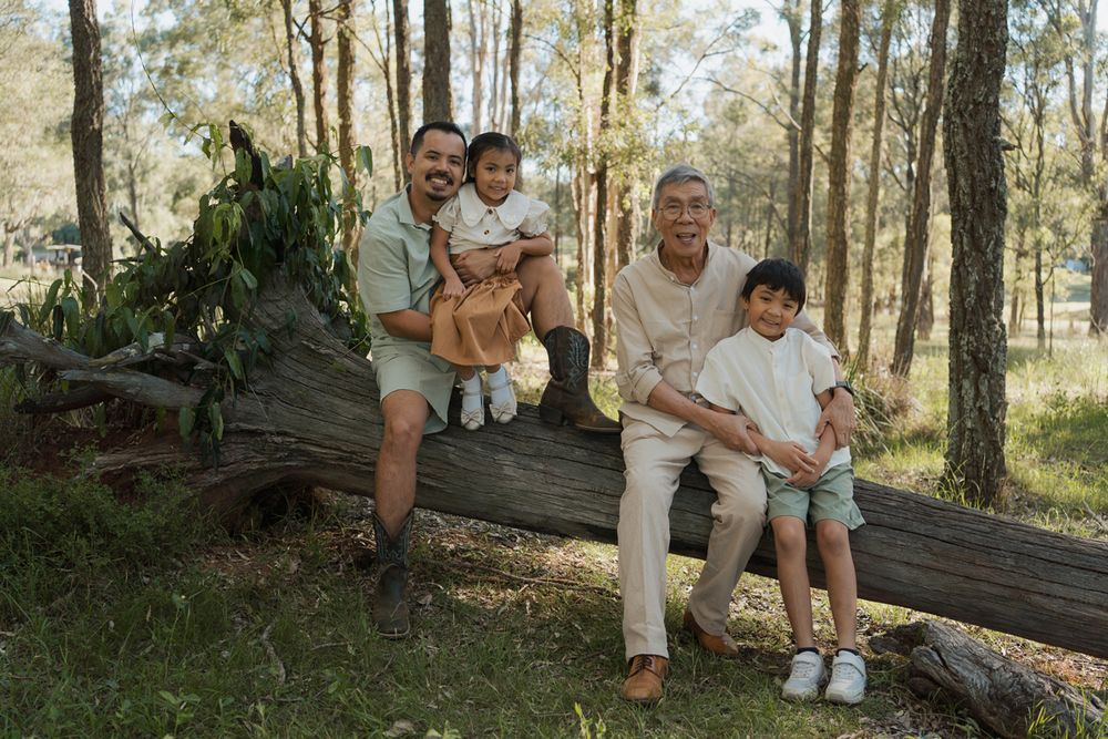 Two men and two children sit on a fallen log in a forest, smiling for a family portrait outdoors in Rouse hill