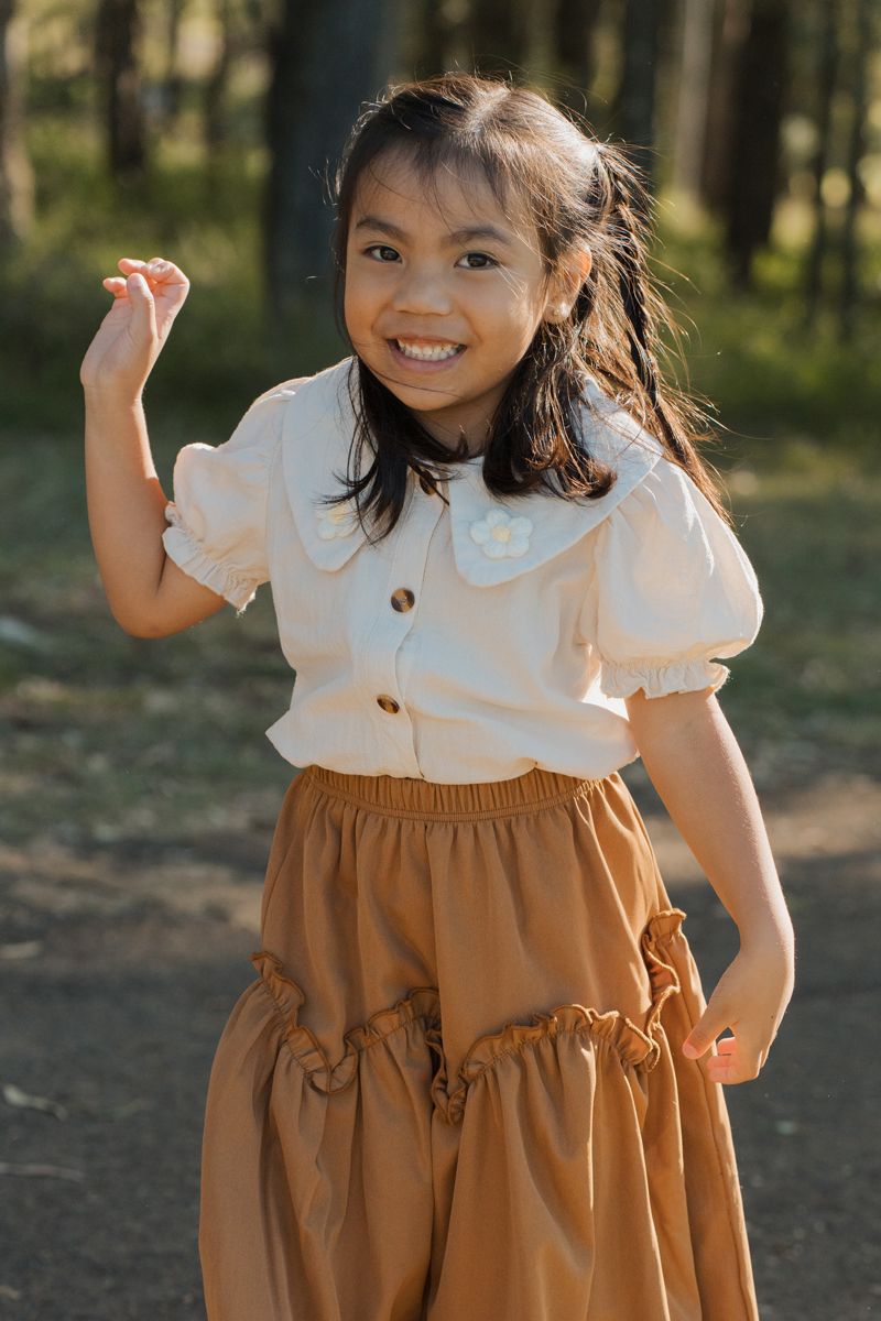 A smiling young girl in a white blouse and brown ruffled skirt poses outdoors in a sunlit forest setting. During a family photoshoot in Sydn