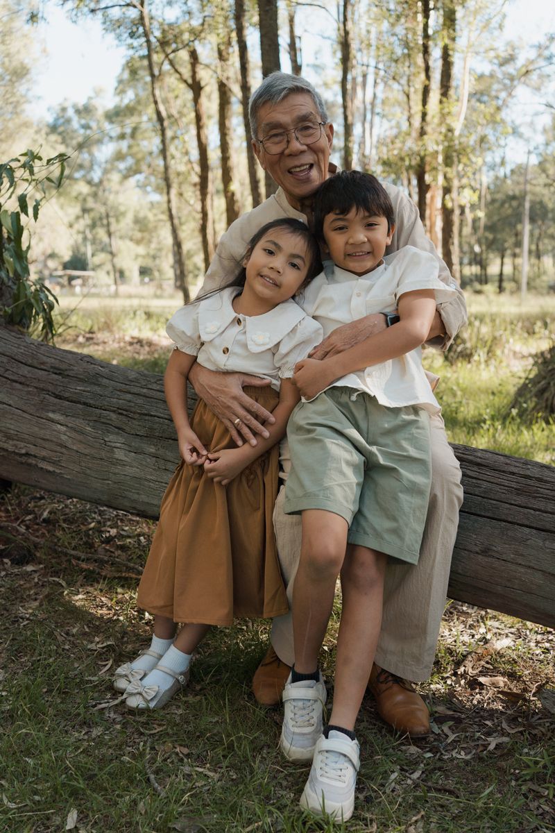 Grandfather hugging two young grandchildren while leaning on a log in a sunny outdoor Rouse hill regional park family photoshoot